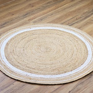 Peut inclure: Un tapis rond en jute de couleur beige naturel avec une bordure blanche. Le tapis est fait de fibres tiss&eacute;es et est pos&eacute; sur un sol en bois. Le diam&egrave;tre du tapis est d'environ 120 cm.