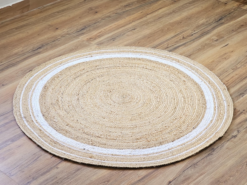 Peut inclure: Un tapis rond en jute de couleur beige naturel avec une bordure blanche. Le tapis est fait de fibres tiss&eacute;es et est pos&eacute; sur un sol en bois. Le diam&egrave;tre du tapis est d'environ 120 cm.