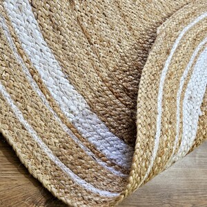 Peut inclure: Un tapis rond en jute avec un motif tiss&eacute;. Le tapis pr&eacute;sente des bandes altern&eacute;es de beige naturel et de blanc, cr&eacute;ant un motif ray&eacute;. Le tapis est partiellement enroul&eacute;, r&eacute;v&eacute;lant son envers.