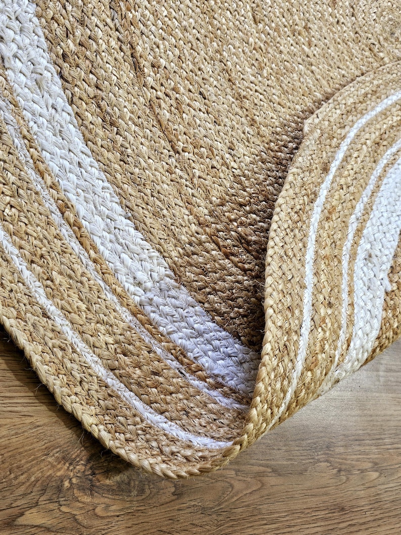 Peut inclure: Un tapis rond en jute avec un motif tiss&eacute;. Le tapis pr&eacute;sente des bandes altern&eacute;es de beige naturel et de blanc, cr&eacute;ant un motif ray&eacute;. Le tapis est partiellement enroul&eacute;, r&eacute;v&eacute;lant son envers.