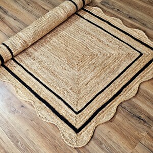 May include: A rolled-up, rectangular jute rug with a scalloped edge. The rug features a natural tan color with a black border and a black stripe on the rolled portion. The rug is placed on a wooden floor.