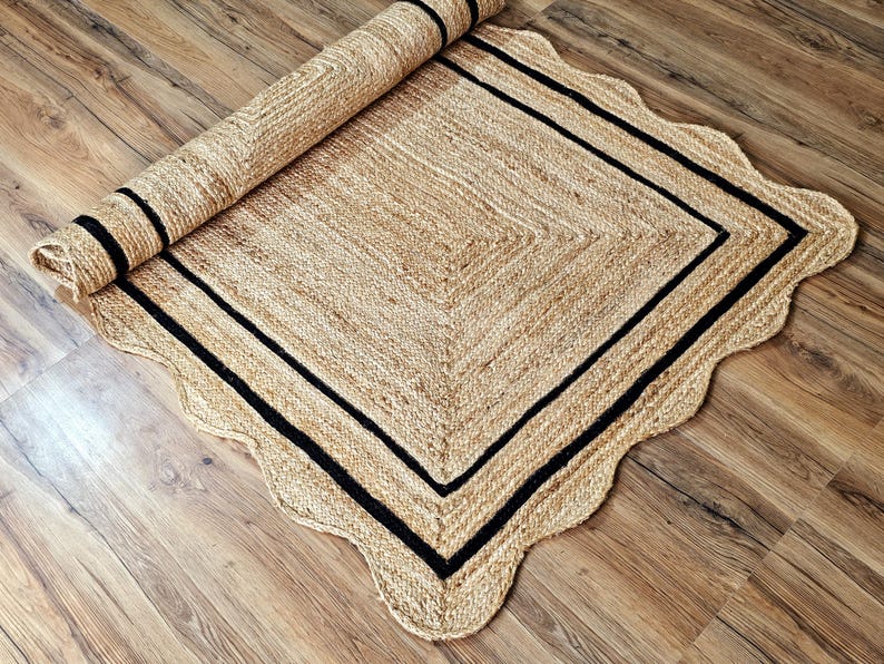 May include: A rolled-up, rectangular jute rug with a scalloped edge. The rug features a natural tan color with a black border and a black stripe on the rolled portion. The rug is placed on a wooden floor.