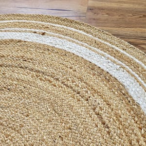 Peut inclure: Gros plan sur un tapis rond en jute de couleur beige naturel avec un motif &agrave; rayures blanches. Le tapis est fait de fibres tiss&eacute;es et a une surface textur&eacute;e. Le tapis est sur un sol en bois.
