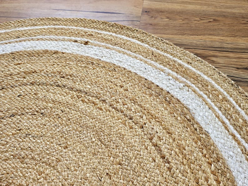 Peut inclure: Gros plan sur un tapis rond en jute de couleur beige naturel avec un motif &agrave; rayures blanches. Le tapis est fait de fibres tiss&eacute;es et a une surface textur&eacute;e. Le tapis est sur un sol en bois.