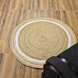 Peut inclure: Tapis rond en jute avec un centre beige naturel et une bordure blanche. Le tapis est pos&eacute; sur un sol en bois, avec un fauteuil gris fonc&eacute; au premier plan. La texture et le design du tapis ajoutent une touche rustique.