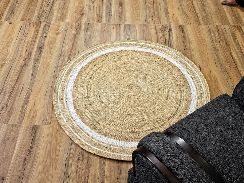 Peut inclure: Tapis rond en jute avec un centre beige naturel et une bordure blanche. Le tapis est pos&eacute; sur un sol en bois, avec un fauteuil gris fonc&eacute; au premier plan. La texture et le design du tapis ajoutent une touche rustique.