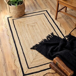 May include: A rectangular jute rug with a scalloped edge and a black border. A black throw blanket with fringe, a woven basket with a plant, and two vintage books are on the wooden floor. A small wooden table with a mug is in the background.