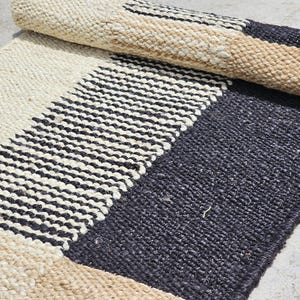 May include: A rolled-up, rectangular rug with a woven design. The rug features sections of off-white, black and white striped, tan, and dark blue. The rug is made of a textured material, likely natural fibers, and is partially rolled up.