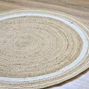 Peut inclure: Un tapis rond en jute de couleur beige naturel, bord&eacute; d'une bordure blanche. Le tapis est fait de fibres tiss&eacute;es et pr&eacute;sente un motif en spirale au centre. Id&eacute;al pour les salons, les chambres ou les entr&eacute;es.