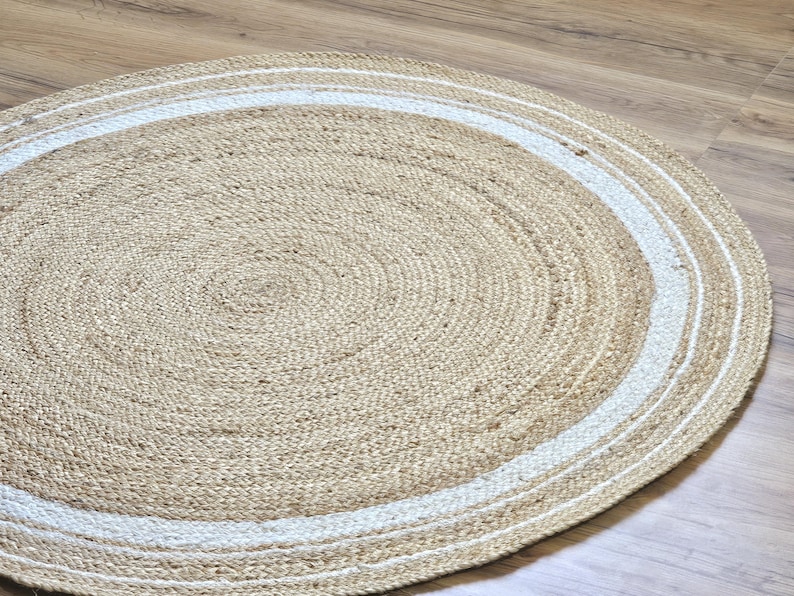 Peut inclure: Un tapis rond en jute de couleur beige naturel, bord&eacute; d'une bordure blanche. Le tapis est fait de fibres tiss&eacute;es et pr&eacute;sente un motif en spirale au centre. Id&eacute;al pour les salons, les chambres ou les entr&eacute;es.