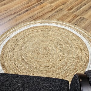 Peut inclure: Un tapis rond en jute de couleur beige naturel avec une bordure blanche. Il est pos&eacute; sur un sol en bois, avec une partie d'une chaise rembourr&eacute;e gris fonc&eacute; visible. Le tapis pr&eacute;sente un motif de cercles concentriques.