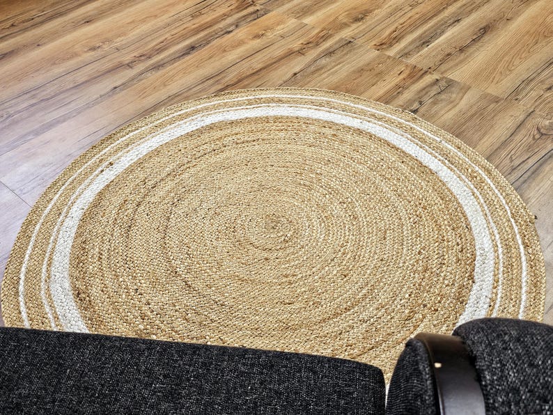 Peut inclure: Un tapis rond en jute de couleur beige naturel avec une bordure blanche. Il est pos&eacute; sur un sol en bois, avec une partie d'une chaise rembourr&eacute;e gris fonc&eacute; visible. Le tapis pr&eacute;sente un motif de cercles concentriques.