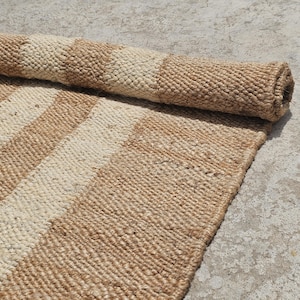 May include: A rolled-up, rectangular rug with alternating beige and light brown stripes. The rug is made of a woven, textured material, and is partially unrolled, revealing the striped pattern. The rug is on a light gray surface.