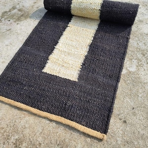 Puede incluir: Una alfombra rayada en blanco y negro con un borde beige. La alfombra está hecha de fibras naturales y tiene una textura tejida.