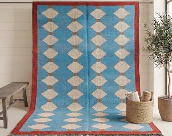 Puede incluir: Una alfombra rectangular que presenta un campo azul brillante con un patrón repetitivo de diamantes blancos conectados por líneas marrones finas. La alfombra está enmarcada por un borde rojo sólido y terminada con flecos gris oscuro.