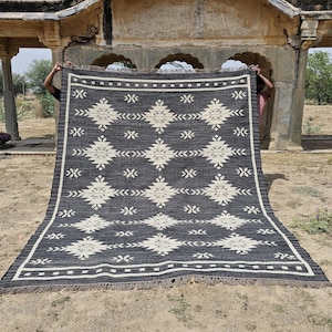 May include: A black and white geometric patterned rug with fringe. The rug is being held up by two people in front of a stone archway.