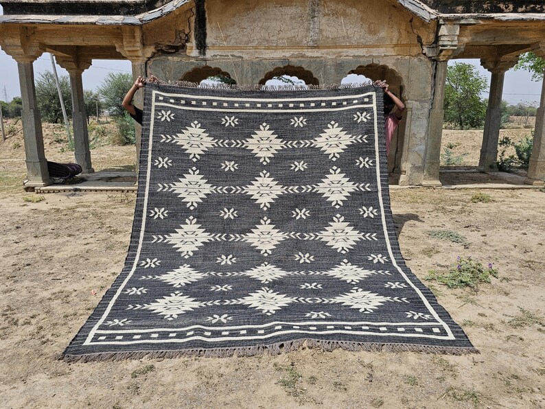 May include: A black and white geometric patterned rug with fringe. The rug is being held up by two people in front of a stone archway.