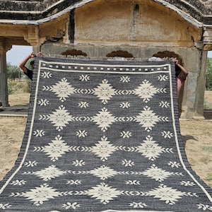 May include: A black and white geometric patterned rug with a fringe border. The rug is being held up by two people in front of a stone archway.