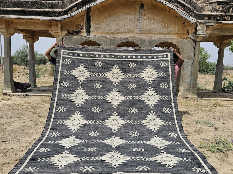 May include: A black and white geometric patterned rug with a fringe border. The rug is being held up by two people in front of a stone archway.