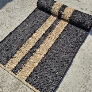 Puede incluir: Una alfombra negra y de yute natural con dos rayas anchas que recorren la longitud de la alfombra.