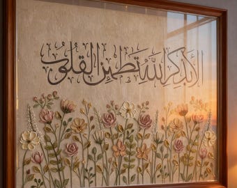 Op de afbeelding: Groot ingelijst kunstwerk met Arabische kalligrafie en bloemenreliëf. Het kunstwerk staat in een donkere houten lijst en heeft een beige achtergrond met een bloemstuk van roze, witte en perzikbloemen. Een gebedskleed en kaarsen staan op de voorgrond.