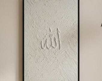 May include: Textured white wall art with a black frame. The artwork features the Arabic word "Allah" in a raised, textured design. The piece is displayed on a neutral-colored wall, complemented by a black pendant light and a decorative towel rack.