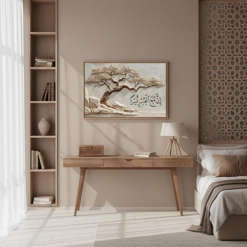 May include: A framed relief sculpture of a tree with Arabic calligraphy. The artwork is in neutral tones and hangs above a wooden desk. A lamp, books, and decorative box are on the desk. A bookshelf and bed are also visible.