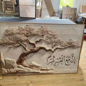 May include: A framed, textured relief artwork featuring a detailed tree with a sprawling trunk and branches, and a light brown color. The artwork includes Arabic calligraphy. The piece is set against a light background.