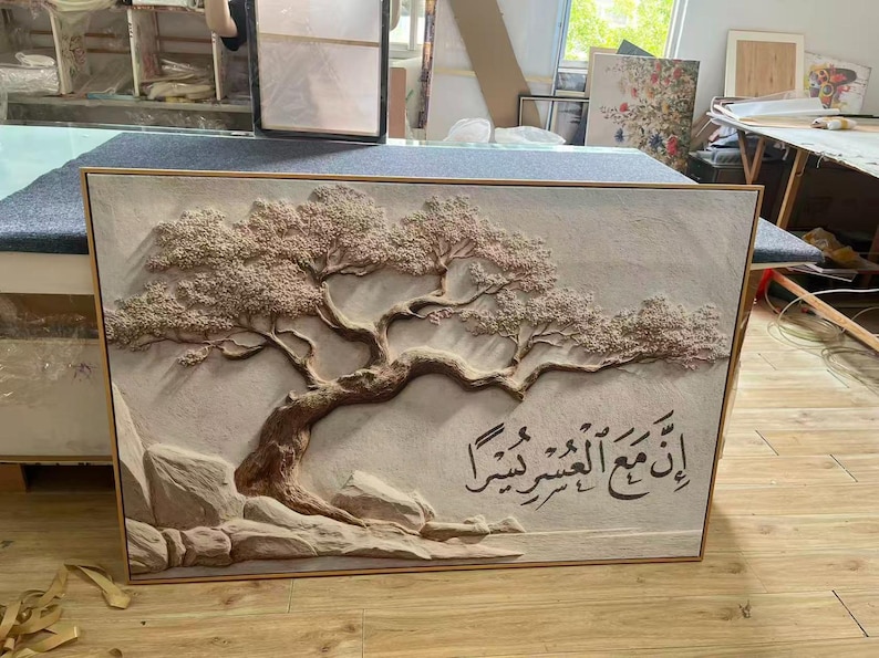 May include: A framed, textured relief artwork featuring a detailed tree with a sprawling trunk and branches, and a light brown color. The artwork includes Arabic calligraphy. The piece is set against a light background.