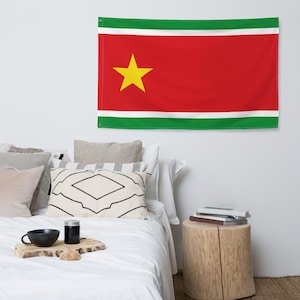 Peut inclure: Un drapeau rectangulaire représentant le drapeau national du Suriname. Le drapeau a un champ rouge avec une étoile jaune au centre. Des bandes horizontales vertes et blanches bordent les bords supérieur et inférieur. Le drapeau est accroché à un mur blanc.
