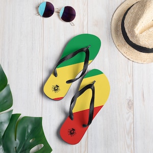 Puede incluir: Un par de chanclas negras con un diseño verde, amarillo y rojo. Las chanclas tienen un logotipo de cabeza de león con el texto "Rasta Vibes" en las correas.
