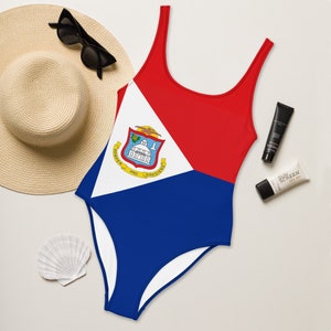 May include: A red, white, and blue swimsuit with the flag of Sint Maarten. The swimsuit has a scoop neck and a low back. The flag is centered on the front of the swimsuit.