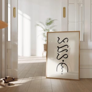 May include: A framed art print with a minimalist design. The artwork features black, abstract lettering and symbols on a cream-colored background, encased in a light wood frame. The print is leaning against a wall in a bright, airy room.