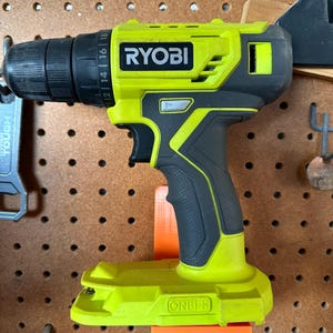 May include: A cordless Ryobi drill, featuring a bright yellow and black design. The drill is mounted on an orange holder against a brown pegboard. The Ryobi logo is prominently displayed. The drill has a black chuck and a variable speed setting.