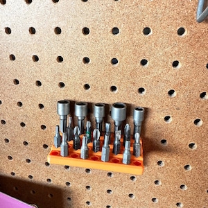 Op de afbeelding: Een assortiment metalen schroevendraaierbits en houders, georganiseerd in een oranje plastic houder. De bits zijn in verschillende maten en vormen, ontworpen voor verschillende schroeftypes. De houder is gemonteerd op een pegboard.
