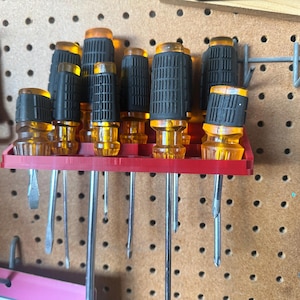 May include: A collection of screwdrivers with clear yellow handles and black grips, stored in a red rack. The tools are arranged on a pegboard, showcasing a variety of sizes and tip styles. A bottle opener is visible on the left.