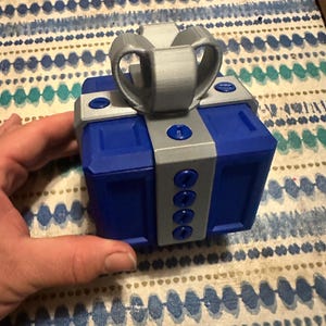 May include: A blue and gray gift box with a bow on top. The box has a square shape with gray accents and a gray bow. The box is held by a hand and sits on a patterned surface.