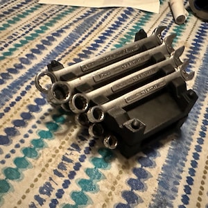 Puede incluir: Un juego de llaves y vasos plateados, cuidadosamente organizados en un soporte negro. Las llaves están etiquetadas con medidas métricas. Las herramientas se muestran sobre una superficie estampada.