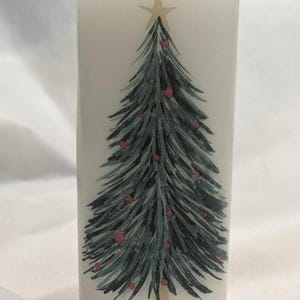 Puede incluir: Vela de pilar blanca con un diseño de árbol de Navidad verde. El árbol tiene una estrella dorada en la parte superior y adornos rosados.