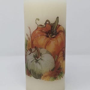 May include: A white pillar candle with a fall-themed design featuring orange and green pumpkins and brown leaves.