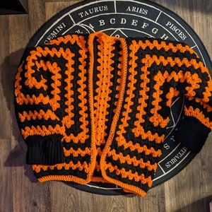Può includere: Un cardigan in crochet nero e arancione con un motivo a righe. Il cardigan è aperto sul davanti e ha maniche lunghe.