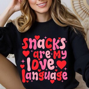 May include: Black sweatshirt with the text "Snacks are my love language" in pink and red. The design includes hearts and stars. The sweatshirt is long-sleeved.