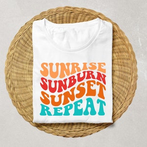 May include: White t-shirt featuring a graphic design that reads "Sunrise Sunburn Sunset Repeat" in a retro font with orange, red, yellow, and blue colours.