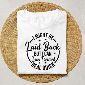 Puede incluir: Camiseta blanca con cuello redondo y el texto negro "I MIGHT BE Laid Back BUT I CAN Lean Forward REAL QUICK" en un círculo. La camiseta está doblada sobre una superficie tejida de color marrón claro.