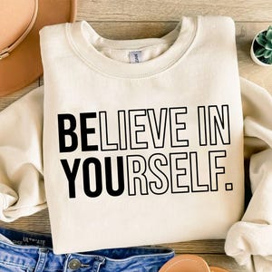 Pode incluir: Camisola creme com o texto "BELIEVE IN YOURSELF" em letras pretas em negrito. A camisola está dobrada, com um chapéu bege e calças de ganga azuis visíveis no fundo. Uma pequena planta suculenta num vaso branco também está presente.