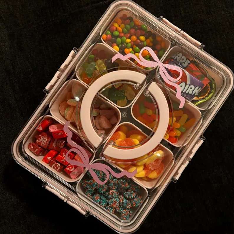 May include: A clear plastic container with multiple compartments filled with different types of candy. The candy includes gummy bears, Skittles, jelly beans, and other assorted candies. The container has a pink lid with a pink handle and pink bows.