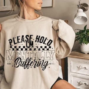 May include: A cream-coloured sweatshirt featuring the black text "PLEASE HOLD I'M MENTALLY Buffering". The text is layered with a chequered pattern and star accents. The sweatshirt has long sleeves.
