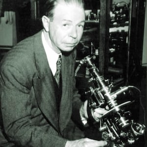 以下が含まれることがあります： スーツを着た男性が顕微鏡を持っている白黒写真。男性はカメラを見ています。顕微鏡は、複数のレンズとダイヤルを備えた複雑な科学機器です。