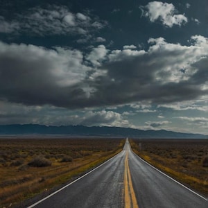 May include: A long, straight tarmac road with yellow lines down the centre, leading to a distant mountain range. The road is surrounded by dry, brown grass and bushes. The sky is overcast with dark clouds.