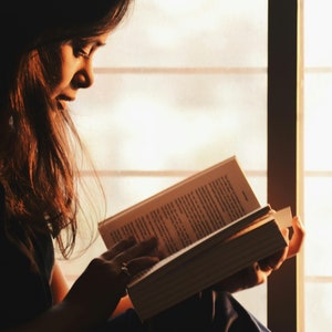 May include: A person sits by a window, reading a book. The light from the window illuminates the pages of the book, casting a warm glow on the person's face.